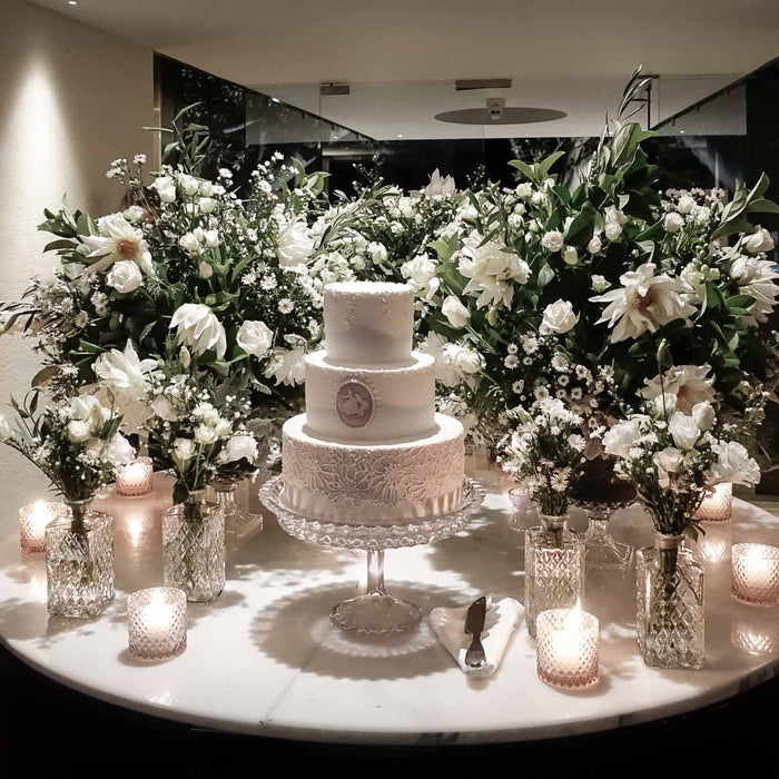 White flowers and foliage behind a tiered wedding cake, with glass candle holders on the table