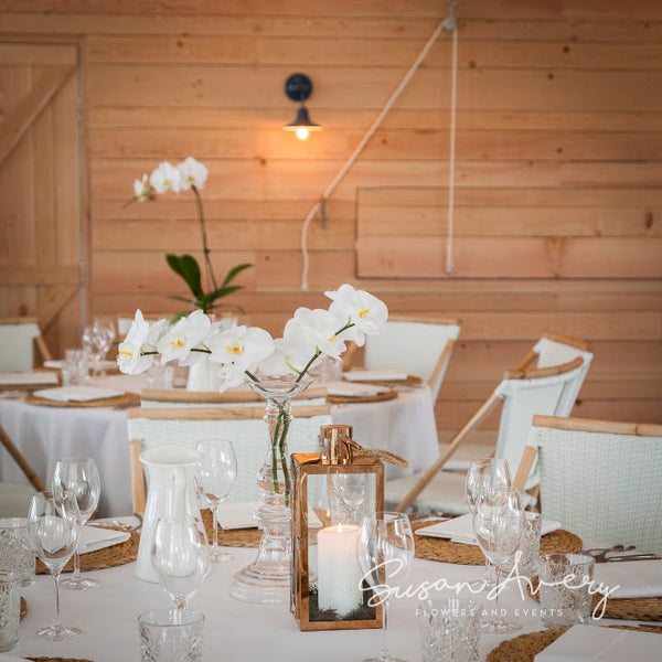 White orchids in clear glass vases with gold lanterns and woven placemats at a Bondi corporate event