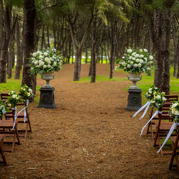 Outdoor ceremony chairs beneath trees with small floral arrangements beside the aisle