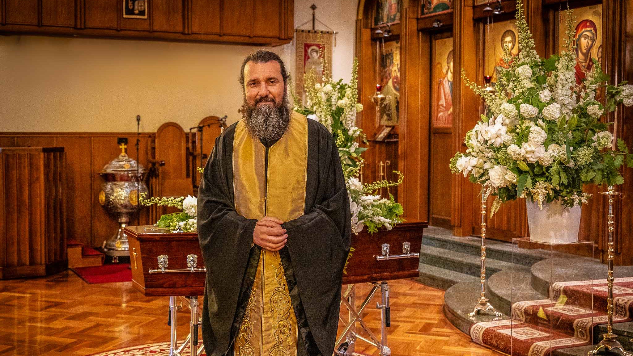 Greek priest in a decorated church interior with floral arrangements and religious icons.
